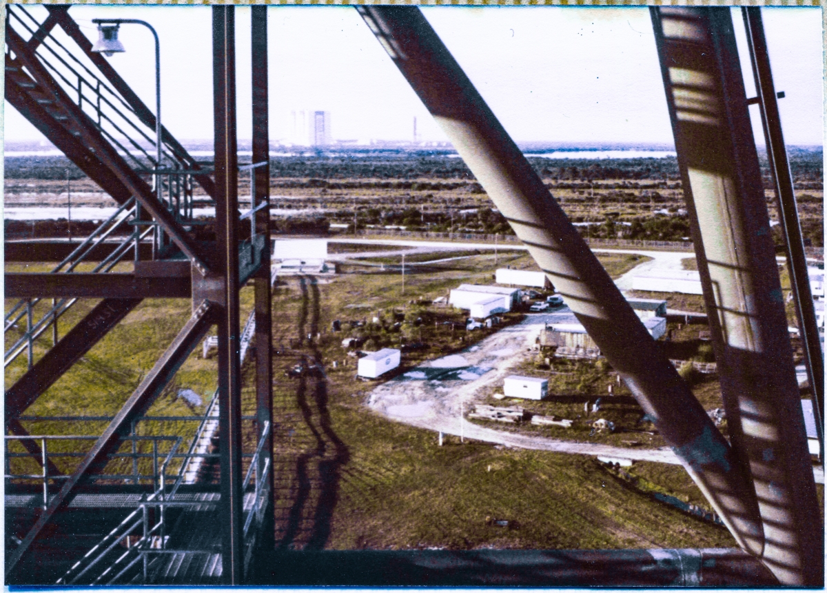 Image 027. From the lowest main platforming level of the Rotating Service Structure at Launch Complex 39-B, Kennedy Space Center, Florida, you are looking south and west toward the Vertical Assembly Building, sitting on the far horizon, tones softened by the haze which comes with such distance. To its right, just beneath the large pipe diagonal of Column Line 7 on the RSS, the small upstanding dark vertical line of the Launch Umbilical Tower (LUT) on Mobile Launcher 1 can be seen. The VAB is where the Saturn V’s were assembled, and ML-1 is the last piece of earth that the crew and the hardware of Apollo 11 touched prior to taking off on their journey to the Moon in July, 1969. In the middle distance between Pad B and the VAB, the stark wetland wilderness of the Merritt Island Launch Area abides. Photo by James MacLaren.